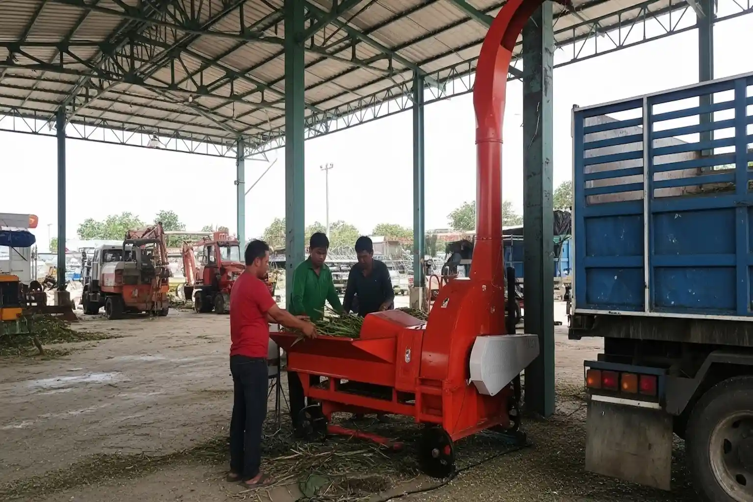 Four men operate a red chaff cutter machine processing crops beside a blue truck.