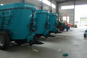 Large teal feed mixer wagons aligned indoors, with tractors and chaff cutters visible behind.