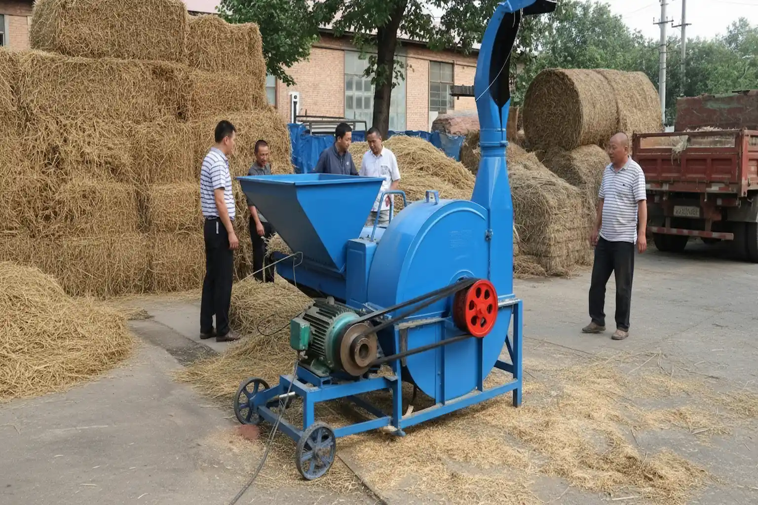 Farmers operating a blue chaff cutter machine surrounded by stacked hay bales outdoors.